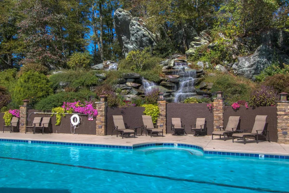 An inviting outdoor swimming pool area bordered by lush landscaping and featuring a cascading waterfall, lounge chairs, and ample space for socializing or sunbathing. The setting is ideal for relaxation during the warmer months.