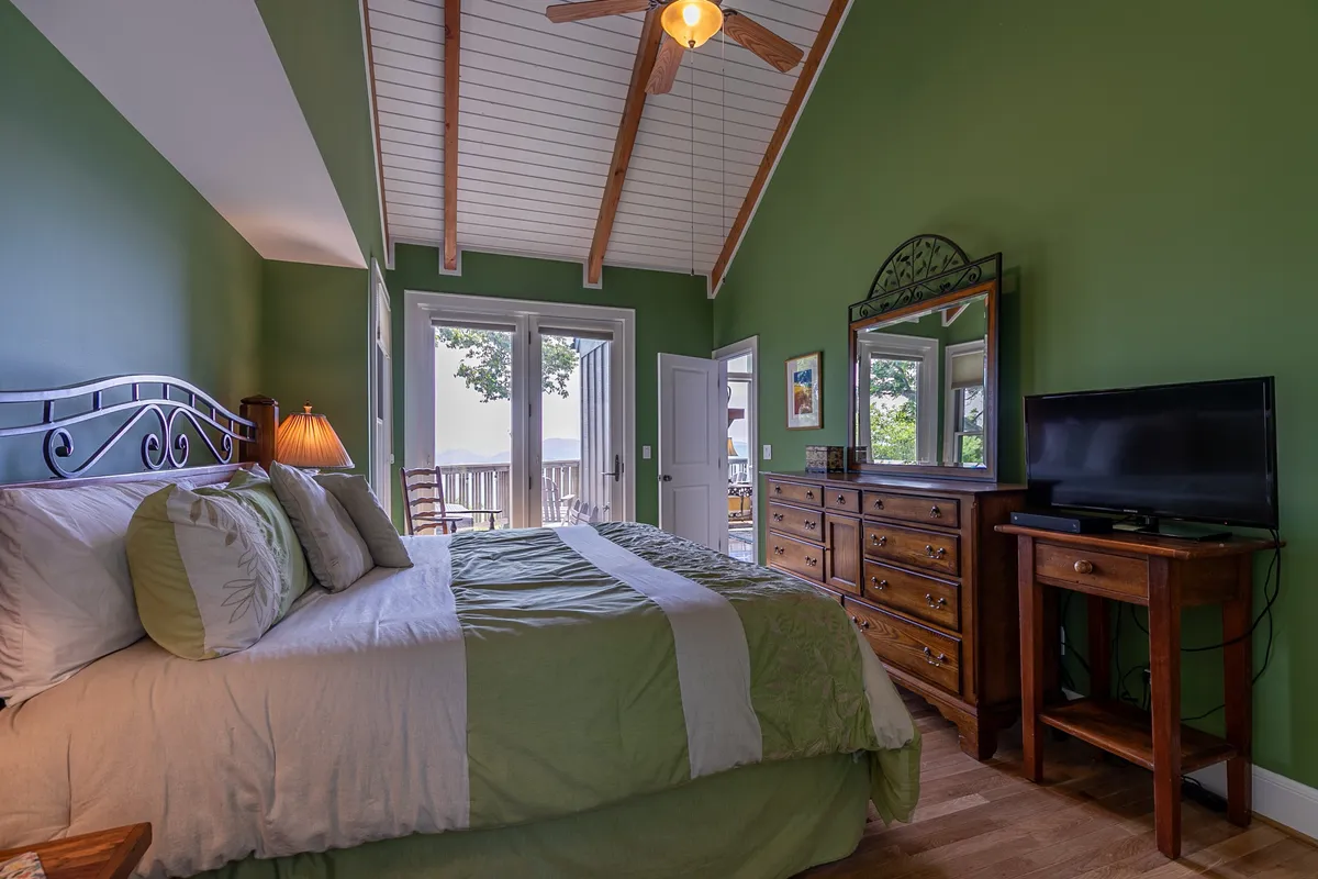 A serene bedroom with a large bed featuring green and white bedding, ample natural light pouring through big windows, a wooden dresser, and a ceiling fan with wooden blades. The room is tranquil and spacious, perfect for restful mornings and relaxed evenings.