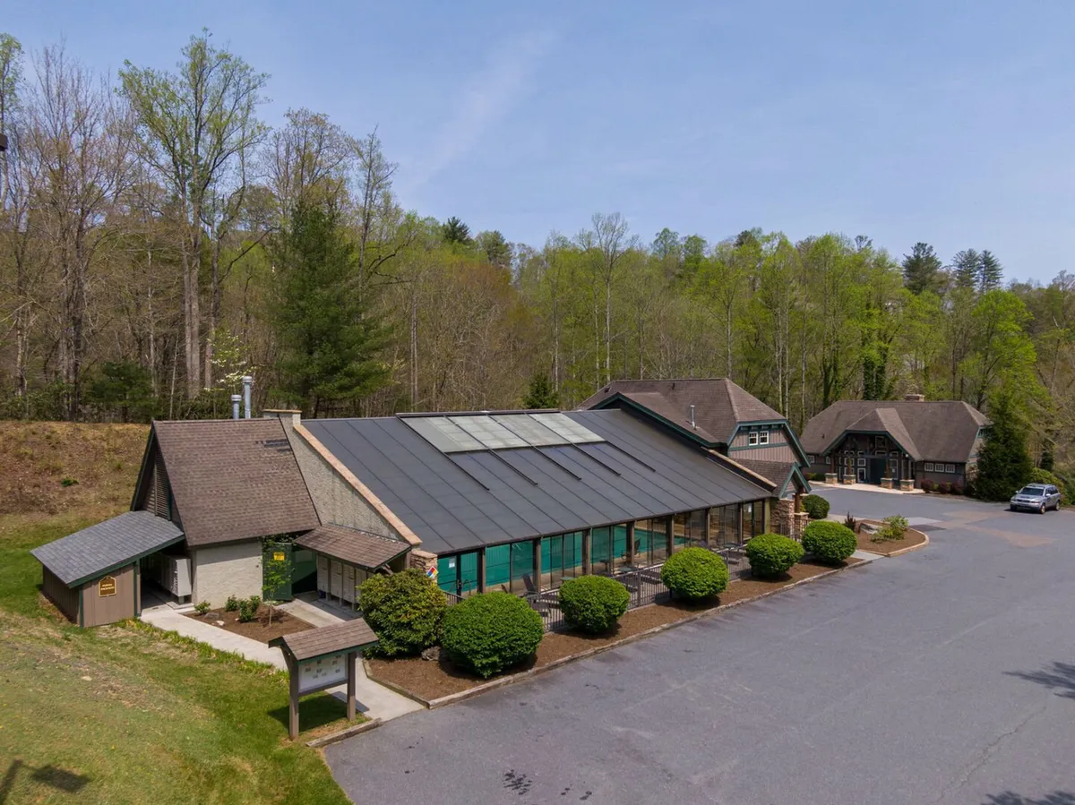 The main building of the retreat is shown nestled among trees and landscaped gardens, with solar panels visible on the roof and a spacious parking area. The setting feels both inviting and environmentally conscious, with the building harmonizing with its surroundings.