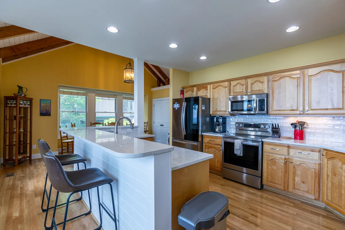 A modern kitchen with a spacious island surrounded by bar stools, stainless steel appliances, warm wooden cabinetry, and large windows letting in natural light. The kitchen opens up to the rest of the living area, making it ideal for socializing and preparing meals together.