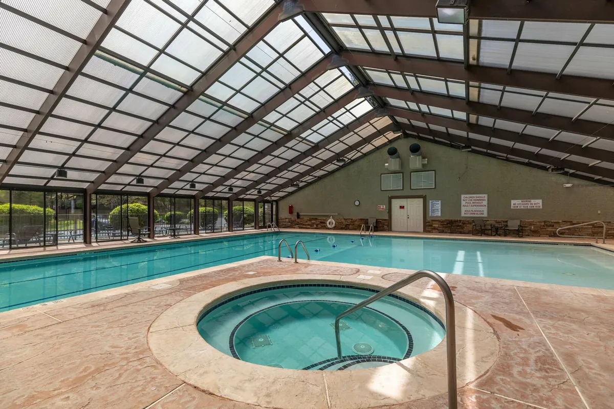A bright, inviting indoor pool area with a large glass window, wooden beams, a hot tub, and lounge chairs. The space exudes serenity and luxury, making it a year-round destination for relaxation and socializing.