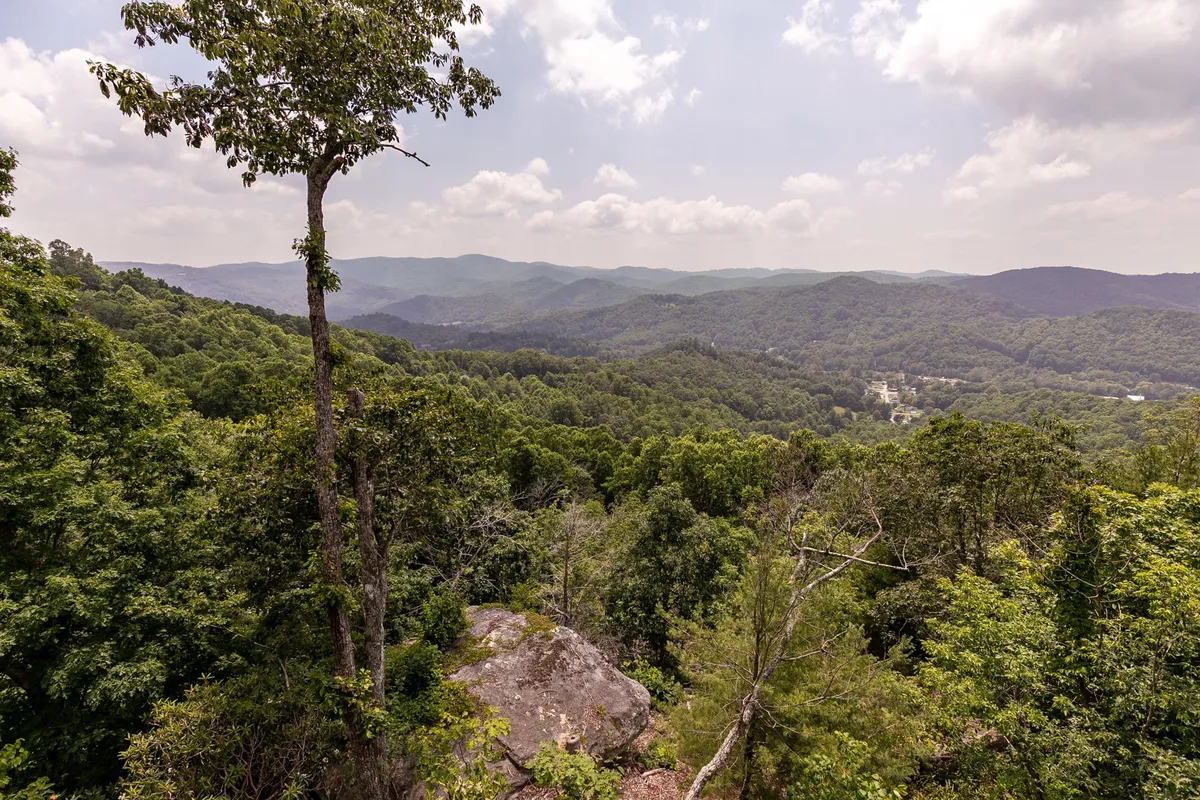 A panoramic view of distant mountains framed by lush greenery and a rugged rocky outcrop in the foreground. The scene radiates tranquility and natural beauty, with clear skies that hint at the range of outdoor adventure possibilities nearby.