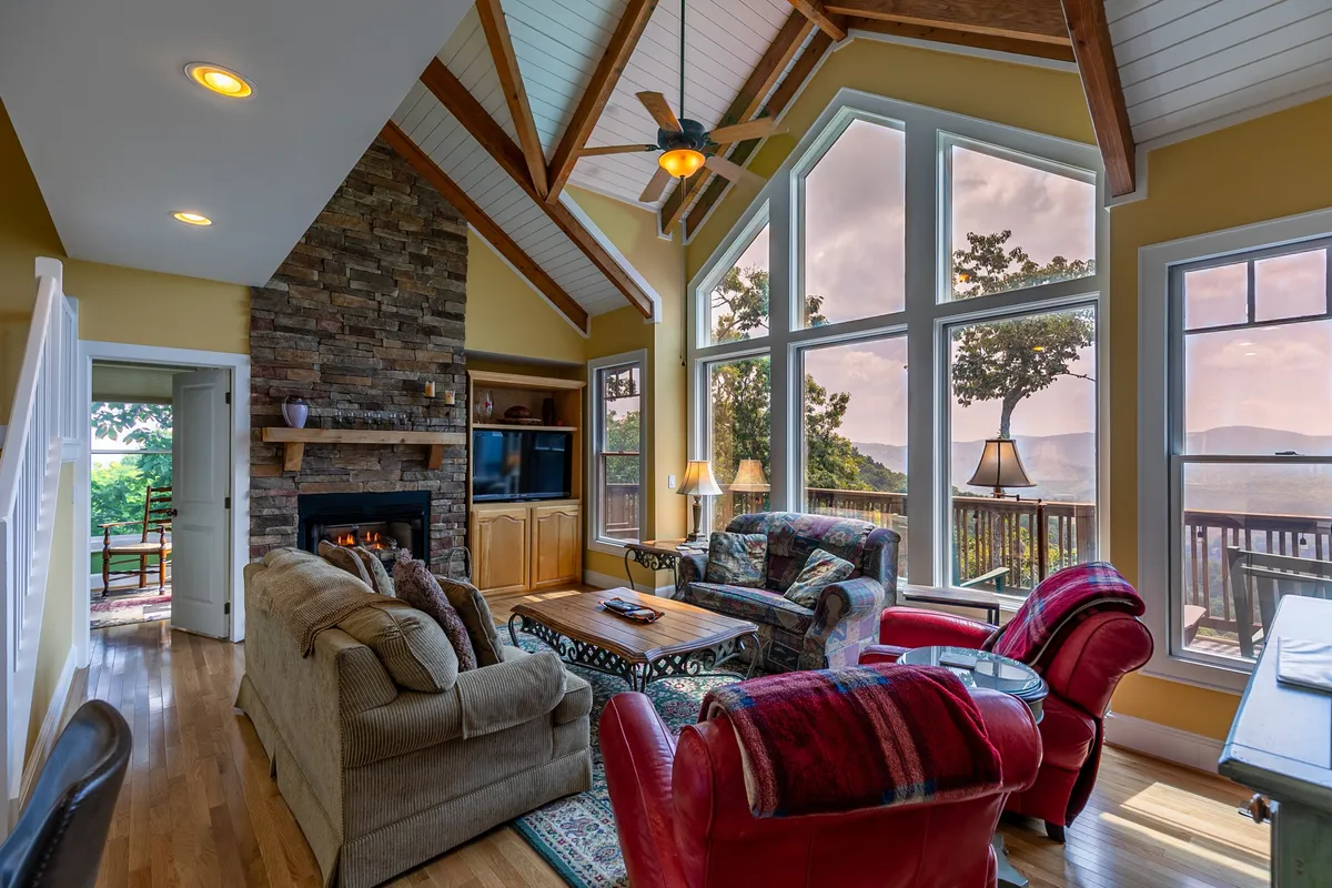 A welcoming open-plan living room featuring large windows that frame the scenic Blue Ridge Mountain views, a stone fireplace, a comfortable seating area, and a dining table designed for family gatherings. The space is bathed in natural light and invites relaxation and conversation.